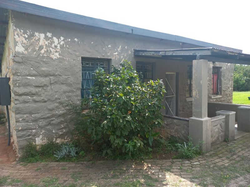 Rear exterior view of a stone-walled house featuring a covered patio area, paved walkway, and security gates, showing signs of significant exterior wear.