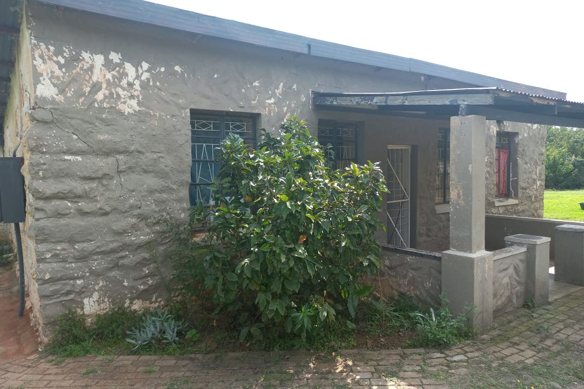 Rear exterior view of a stone-walled house featuring a covered patio area, paved walkway, and security gates, showing signs of significant exterior wear.