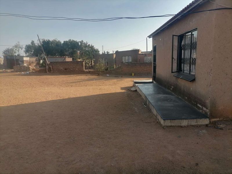 Rear view of the property featuring a concrete patio, security bars on windows, and an expansive unpaved yard area with boundary walling.