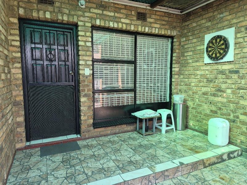A covered patio area featuring exposed brick walls, tiled flooring, and a secure entrance door with an adjacent window.
