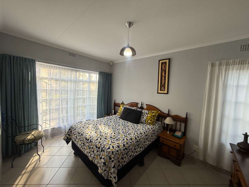 A well-lit bedroom featuring tiled flooring, a central pendant light, and large windows with security bars, offering a functional and comfortable living space.