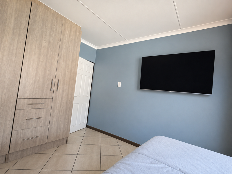 A well-maintained bedroom featuring light wood-grain built-in wardrobes, neutral tiled flooring, and a feature wall with a mounted television.