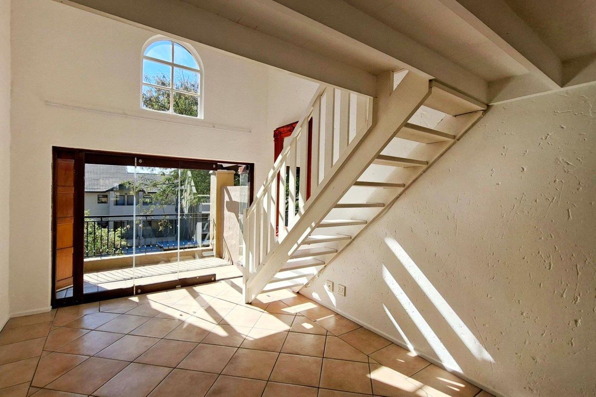 Bright, double-volume living area featuring a staircase, tiled flooring, exposed ceiling beams, and direct access to a balcony through sliding glass doors.