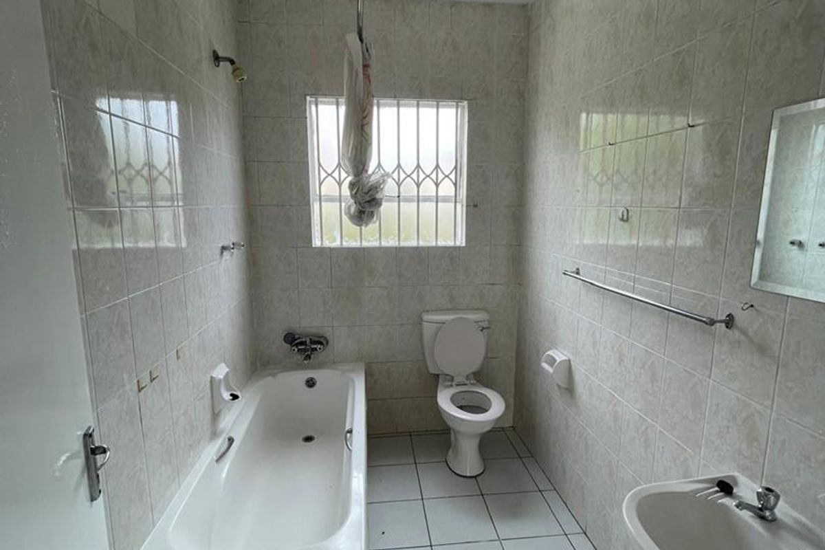 A functional bathroom featuring a built-in bathtub, pedestal basin, and toilet, finished with neutral wall and floor tiles and a security-barred window.