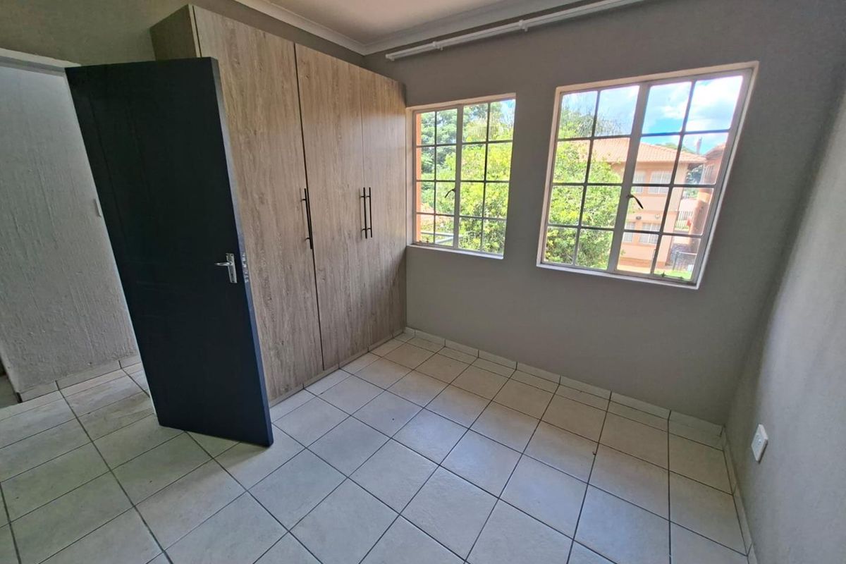 A bright, empty bedroom featuring tiled flooring, built-in wooden cupboards, and large windows providing natural light and a view of the surrounding complex.