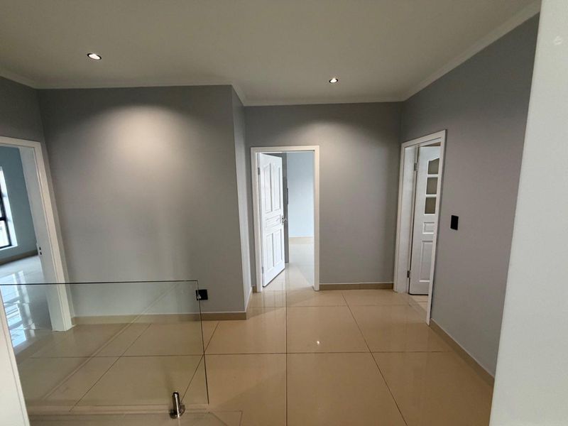A bright, modern hallway featuring neutral grey walls, large format floor tiles, and a contemporary glass balustrade leading to adjacent rooms.