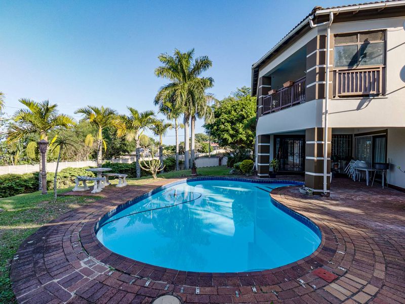 Rear view of the property featuring a swimming pool with brick paving, a landscaped garden, and a covered balcony on the upper level.