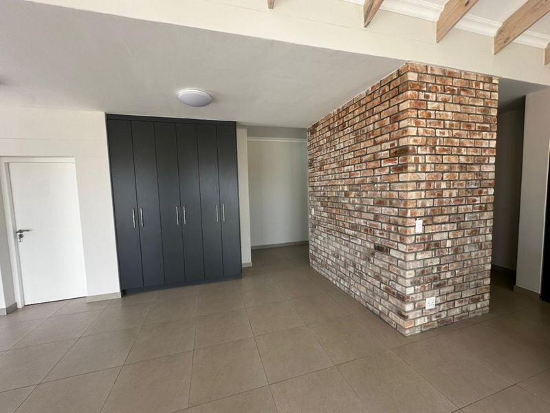 Spacious living area featuring a striking exposed brick feature wall, modern built-in cabinetry, neutral tiled flooring, and exposed timber ceiling beams.