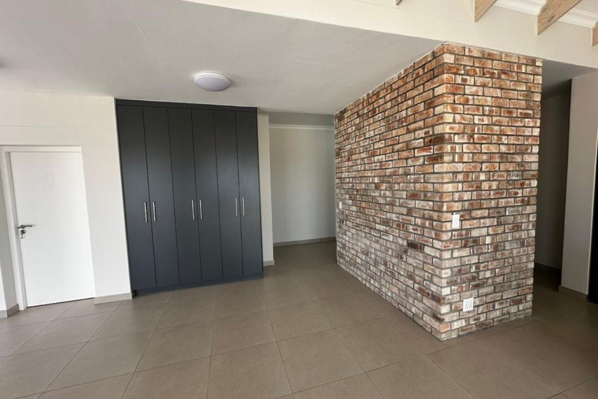 Spacious living area featuring a striking exposed brick feature wall, modern built-in cabinetry, neutral tiled flooring, and exposed timber ceiling beams.