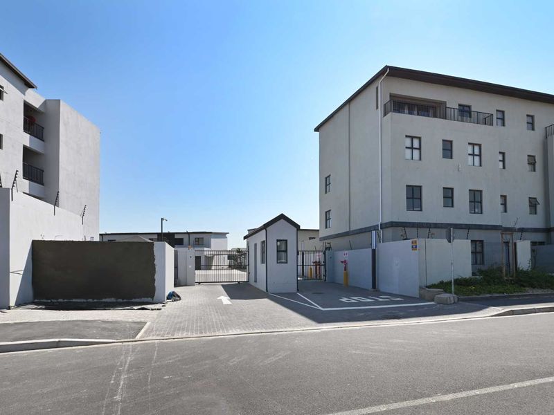 View of the apartment complex entrance featuring a security gate, guard house, and paved driveway with perimeter walling.