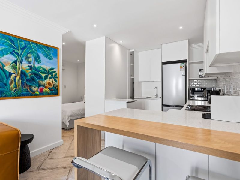 Modern open-plan kitchen featuring sleek white cabinetry, stone countertops, a breakfast bar, and a subway tile backsplash, seamlessly flowing into the adjacent bedroom area.