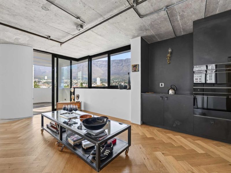 Modern open-plan living area featuring exposed concrete ceilings, herringbone timber floors, sleek black built-in cabinetry, and expansive glass doors with mountain views.