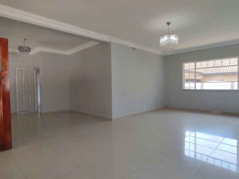 A spacious, bright, and empty living area featuring neutral tiled flooring, crown molding, and a decorative crystal chandelier.