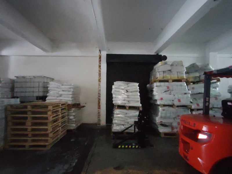 Industrial warehouse storage area featuring exposed concrete beams, a roller shutter door, and concrete flooring, currently utilized for palletized goods storage.