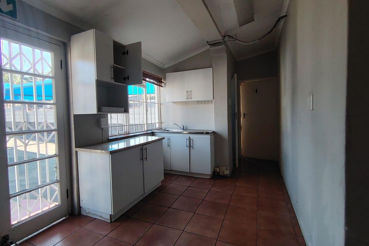 A compact kitchen area featuring built-in white cabinetry, a stainless steel sink, and terracotta-style floor tiles, with direct access to an exterior security door.
