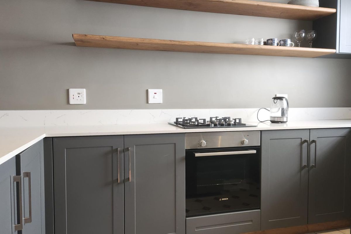 Modern kitchen featuring dark grey shaker-style cabinetry, white quartz countertops, stone splashback, built-in oven, gas hob, and natural timber floating shelves.
