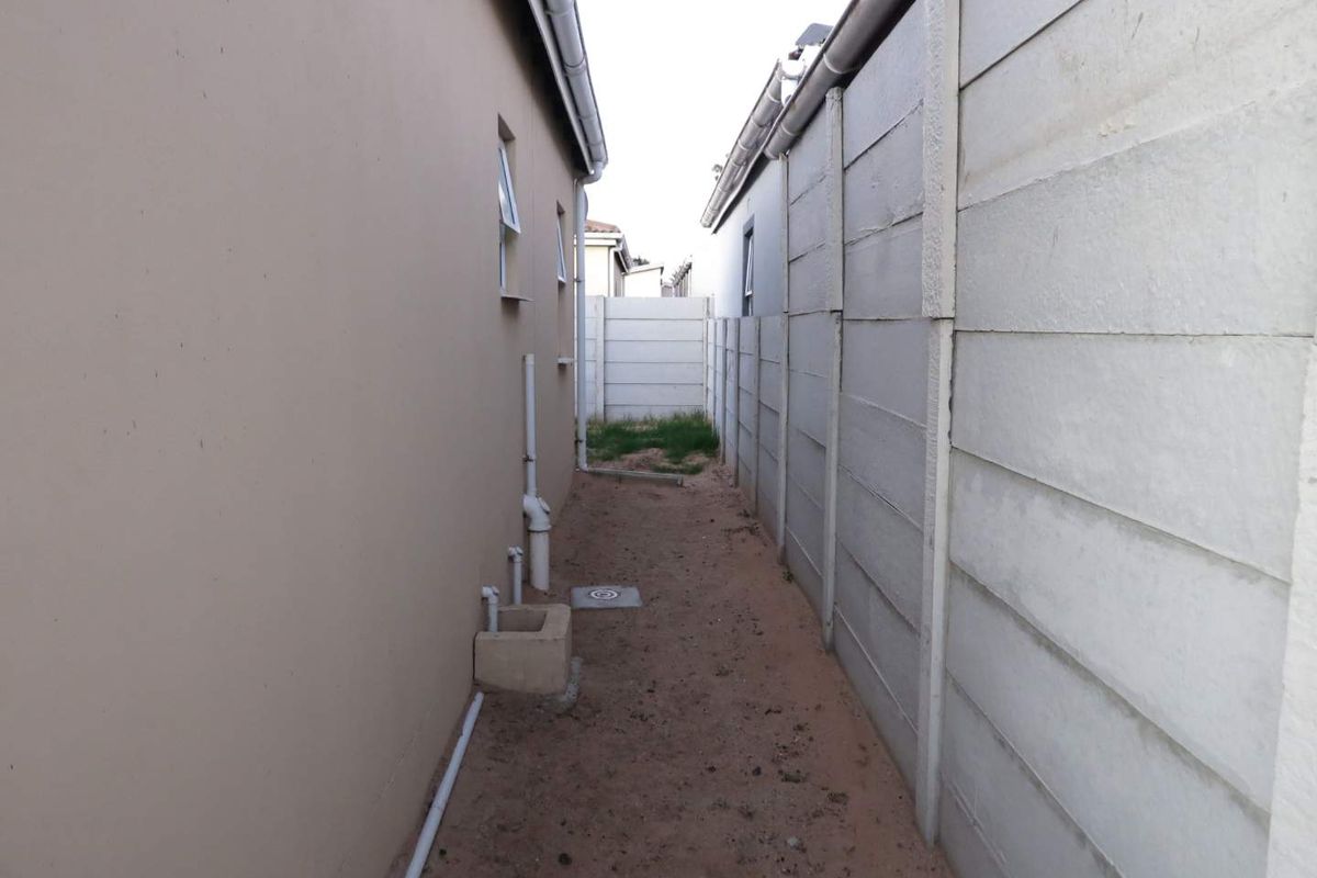 A narrow side passage between the house exterior and a precast concrete boundary wall, featuring exposed plumbing and an unpaved ground surface.