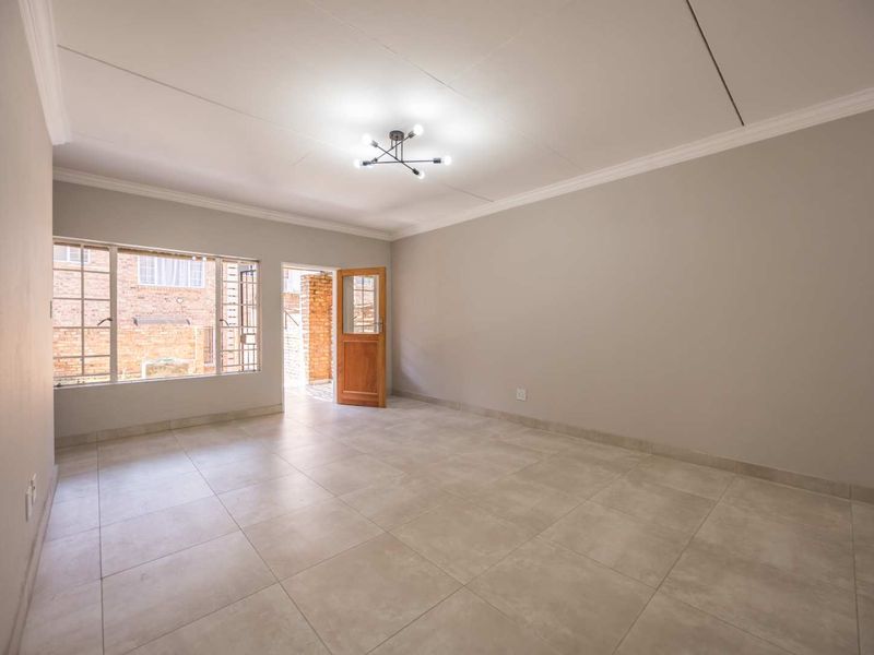 A bright, empty living room featuring neutral tiled flooring, clean walls, ceiling cornicing, and direct access to an outdoor area through a timber door.