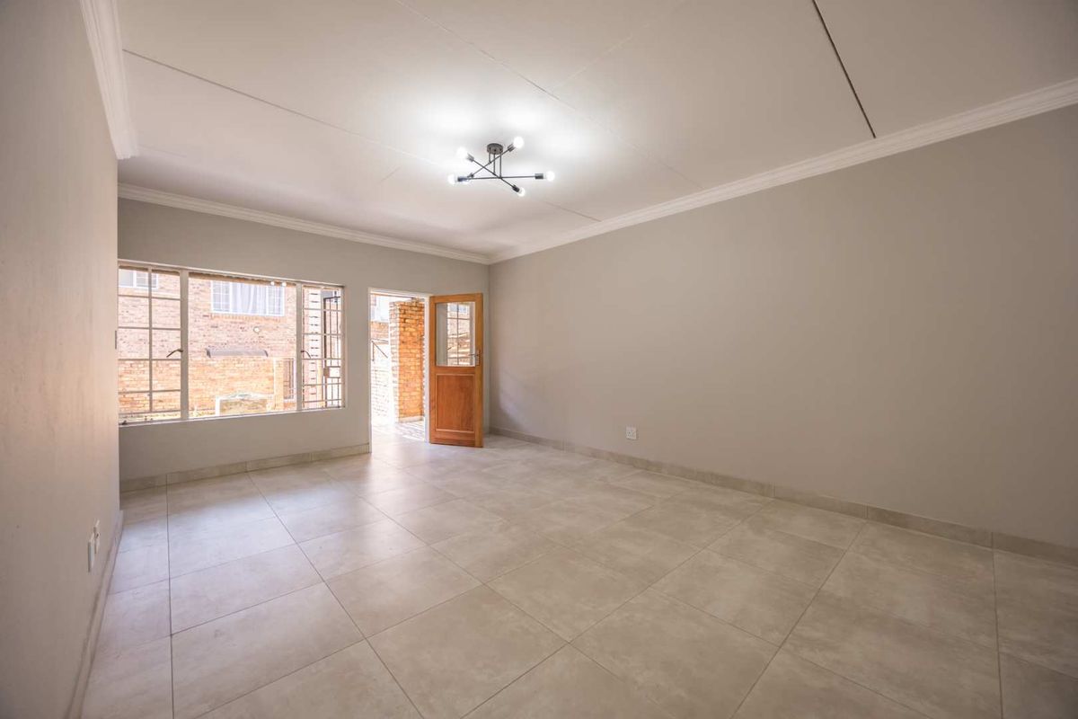 A bright, empty living room featuring neutral tiled flooring, clean walls, ceiling cornicing, and direct access to an outdoor area through a timber door.