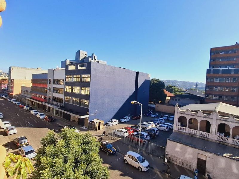An elevated urban view overlooking a street, parking area, and surrounding city buildings under a clear blue sky.