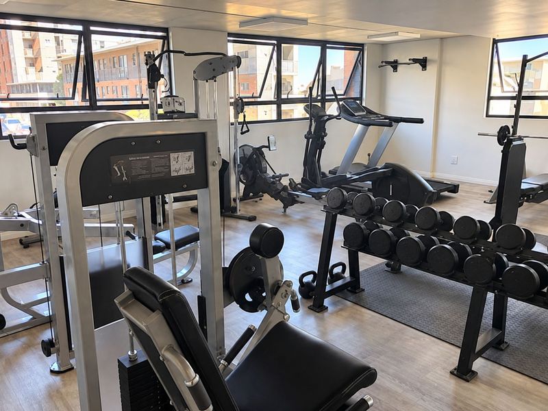 A well-maintained apartment gym facility featuring laminate flooring, large awning windows for natural light, and a variety of fixed fitness equipment.