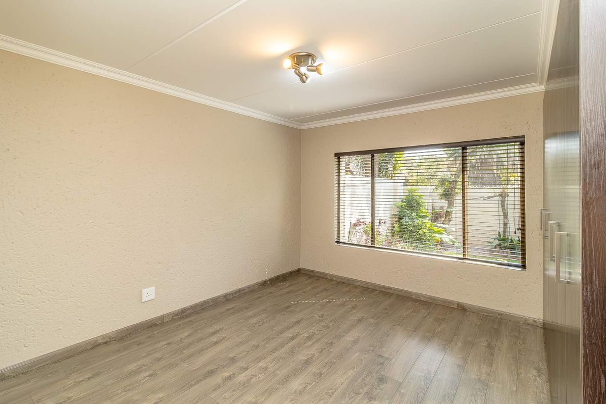 A bright, empty bedroom featuring modern laminate flooring, built-in cupboards, and venetian blinds overlooking the garden.