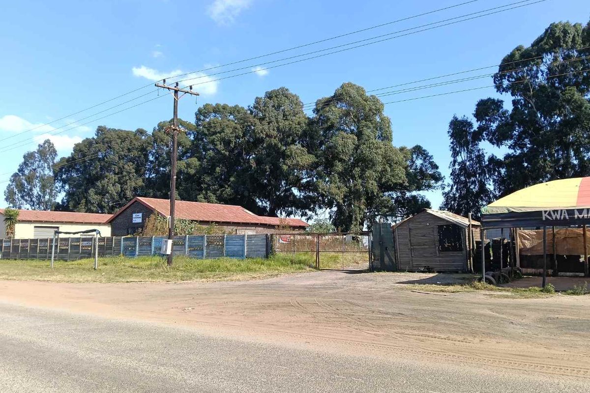 Exterior view of a commercial property featuring a fenced perimeter, gravel access area, and existing structures set against a backdrop of mature trees.