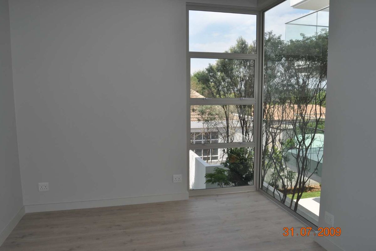A bright, empty bedroom featuring modern light wood-look flooring and large corner windows that provide ample natural light and pleasant outdoor views.
