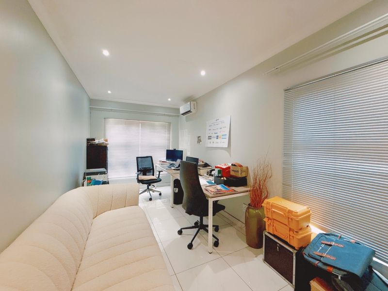 A functional home office featuring tiled flooring, recessed downlighting, a wall-mounted air conditioning unit, and dual windows with fitted venetian blinds.