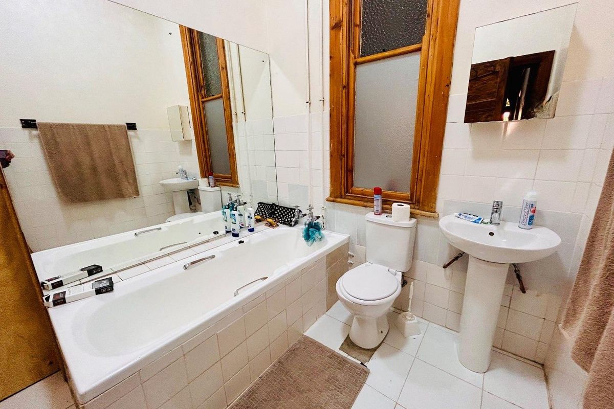A functional bathroom featuring a built-in bathtub, pedestal basin, and toilet, finished with white wall tiles and a wooden window frame.