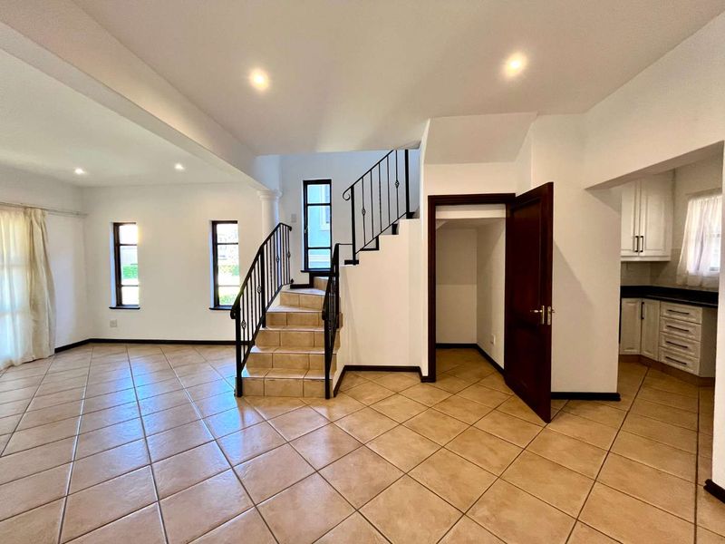 Spacious open-plan living area featuring tiled flooring, a central staircase with wrought iron railing, and an adjacent kitchen with built-in cabinetry.
