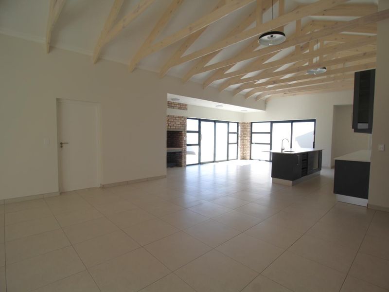 Spacious open-plan living and kitchen area featuring high exposed timber trusses, tiled floors, a built-in brick fireplace, and modern kitchen cabinetry.