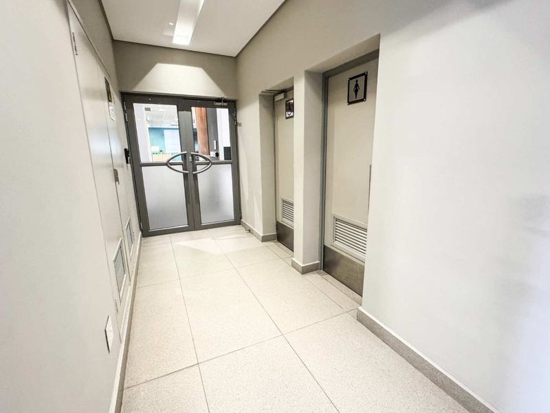 A clean, well-lit commercial hallway featuring tiled flooring, double glass entry doors, and access to restroom facilities.