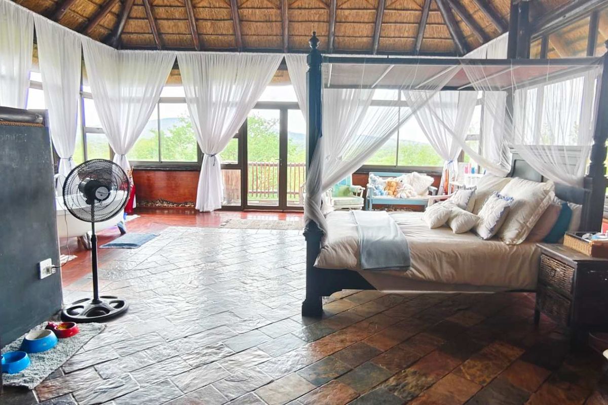 Spacious bedroom featuring a high thatched roof with exposed timber rafters, slate tile flooring, and expansive glass windows offering natural light and scenic views.