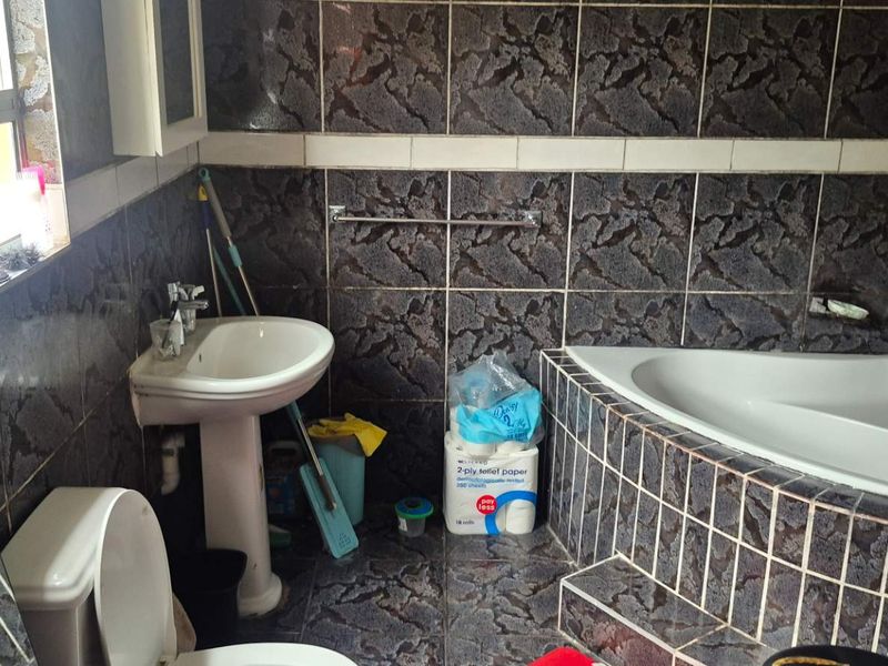 A dated bathroom featuring a corner bathtub, pedestal basin, and toilet with dark patterned wall and floor tiles.