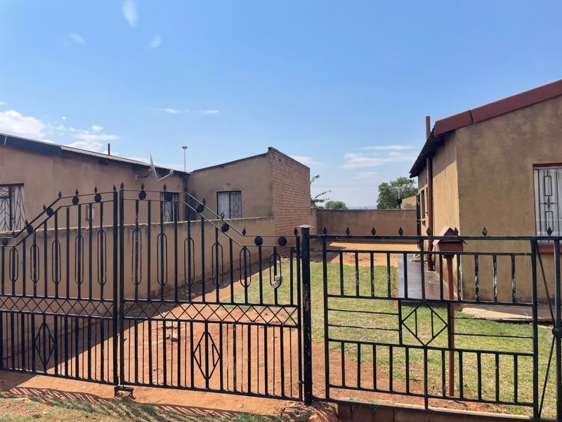 Front exterior view of the property featuring a wrought iron gate, perimeter wall, and basic residential structures with metal roofing.