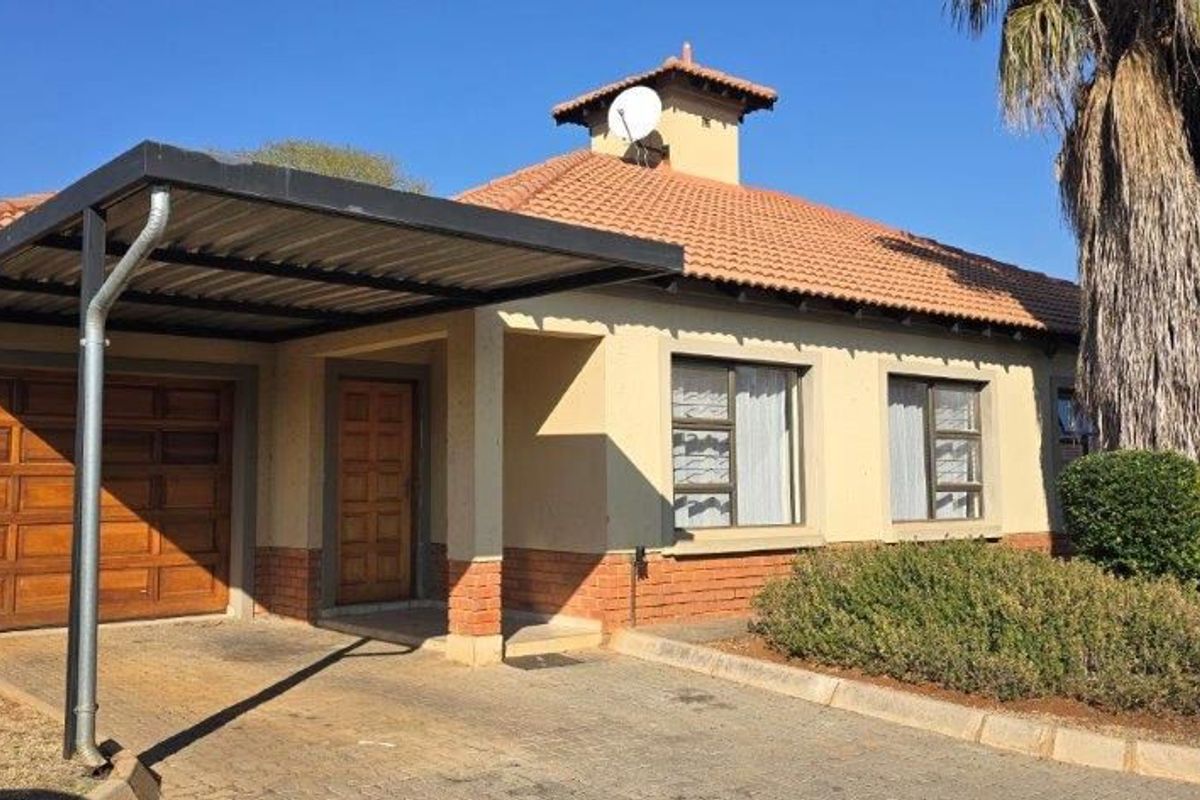 Front elevation of a townhouse featuring a paved driveway, covered carport, wooden garage door, and a tiled roof with established garden landscaping.