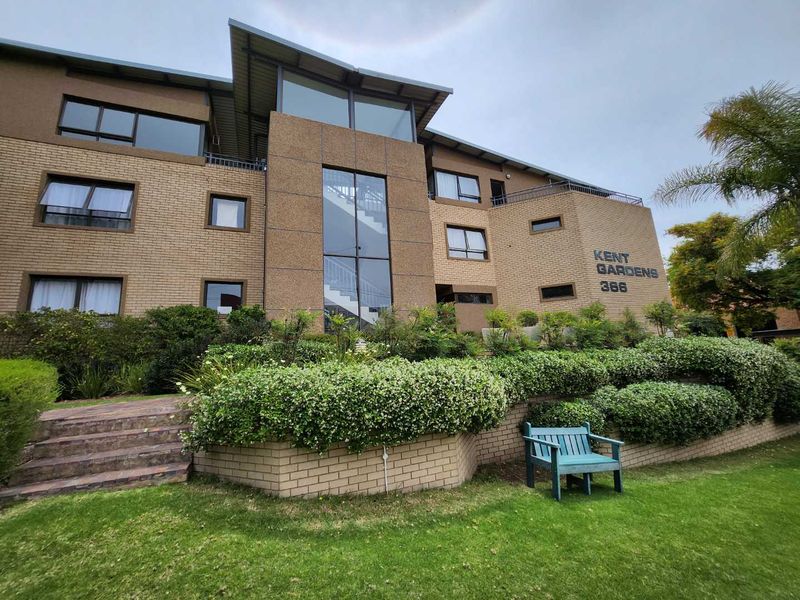 Front elevation of Kent Gardens apartment complex featuring a well-maintained brick facade, large windows, and landscaped garden area with paved access steps.