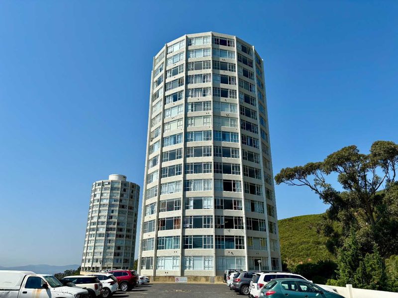 A view of a multi-story residential apartment tower featuring a concrete facade and extensive window glazing, situated above a paved parking area.