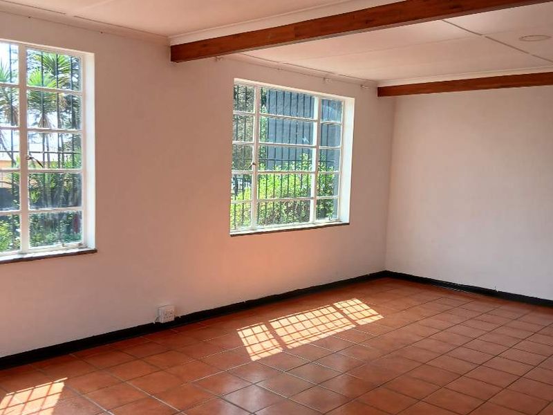 An empty living room featuring terracotta floor tiles, exposed timber ceiling beams, and large steel-framed windows allowing for ample natural light.