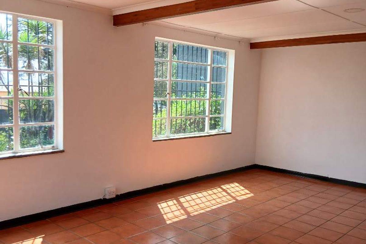 An empty living room featuring terracotta floor tiles, exposed timber ceiling beams, and large steel-framed windows allowing for ample natural light.