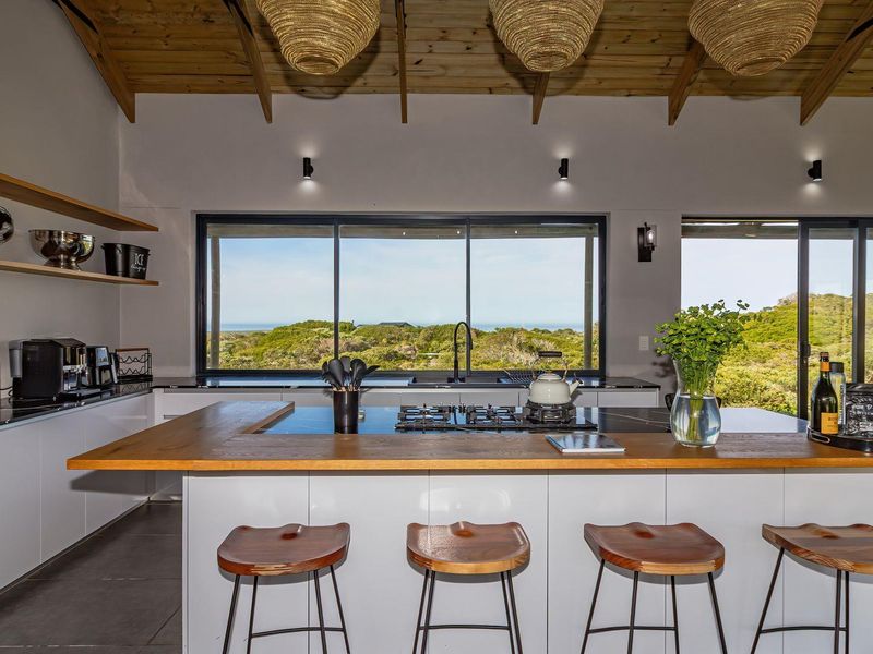 Modern kitchen featuring a large timber-topped island with gas cooktop, exposed beam ceiling, and expansive sliding doors offering scenic coastal views.