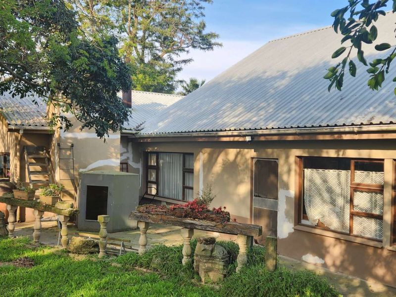 Rear view of a farm-style dwelling featuring a corrugated metal roof, plastered walls, and a stone-balustrade patio area set within a garden.