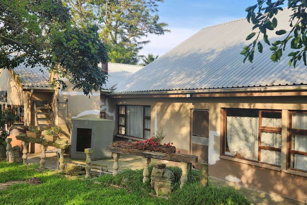 Rear view of a farm-style dwelling featuring a corrugated metal roof, plastered walls, and a stone-balustrade patio area set within a garden.