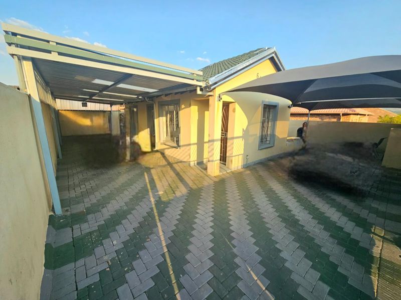 Front exterior view of the house featuring a paved driveway, covered carport, and additional shade sail structure over the entrance area.