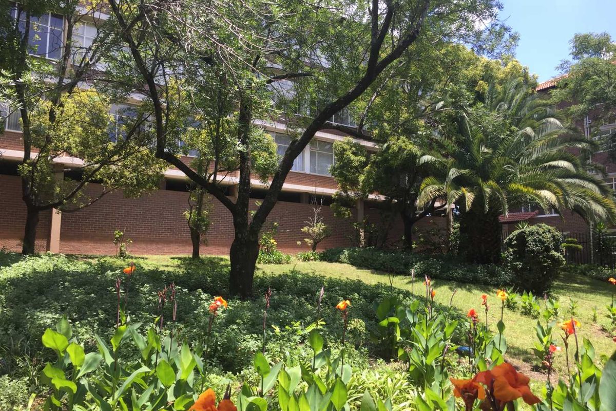 A view of the apartment complex exterior featuring established trees, landscaped garden beds, and brick facade architecture.