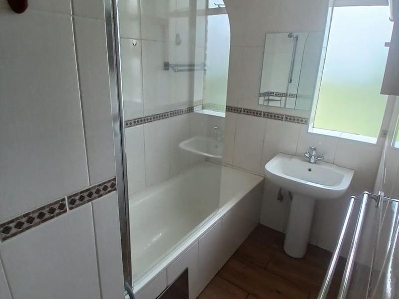 A functional bathroom featuring a bathtub with a glass screen, pedestal sink, and wood-look floor tiles with neutral wall tiling.