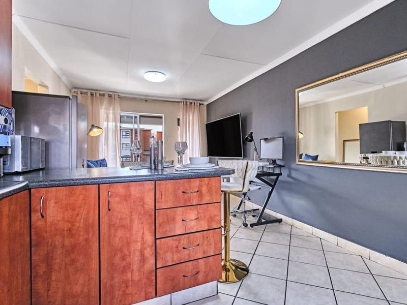 An open-plan apartment living and kitchen area featuring wood-finish cabinetry, dark countertops, ceramic tiled flooring, and a sliding door leading to a balcony.
