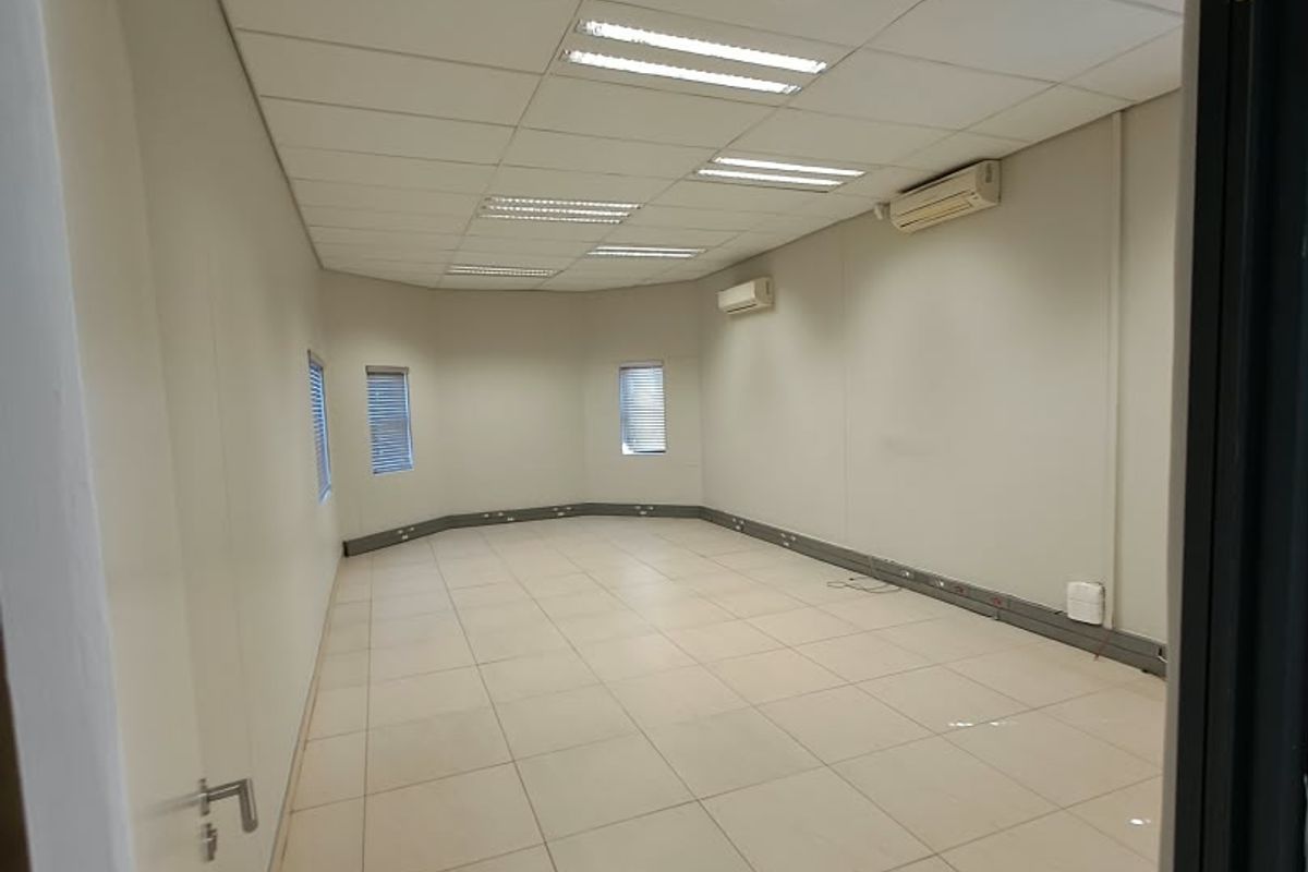 A spacious, empty office area featuring tiled flooring, a suspended ceiling with recessed lighting, and wall-mounted air conditioning units.