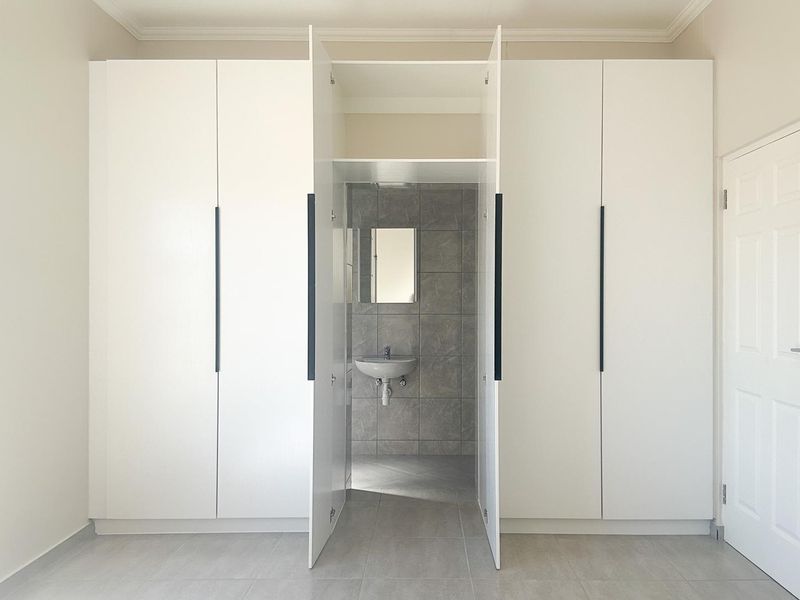 A clean, modern bedroom featuring extensive built-in white cabinetry and a convenient en-suite vanity area with neutral grey tiling.